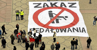 Photograph of a large banner spread on a street. The banner reads ZAKAZ PEDAŁOWANIA. A crowd of people are surrounding, most of them observing the banner.